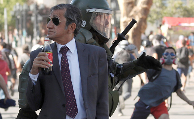 In Dark Glasses, Smartly-Dressed Chilean Sips Soda As Riot Rages Around