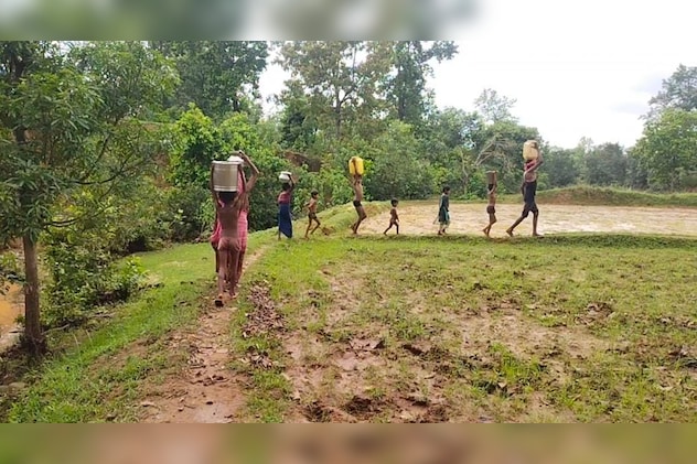 पानी लेकर खेतों की पगडंडी से एक समूह में जाते हुए ग्रामीण और छोटे-छोटे बच्चे. 