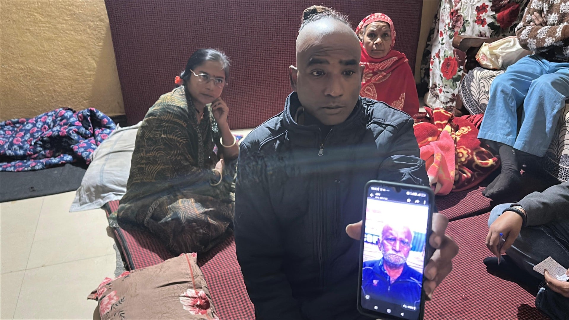 मोबाइल में पिता की तस्वीर दिखाते नंदलाल के पुत्र, पीछे गुमसुम बैठीं घर की महिलाएं.