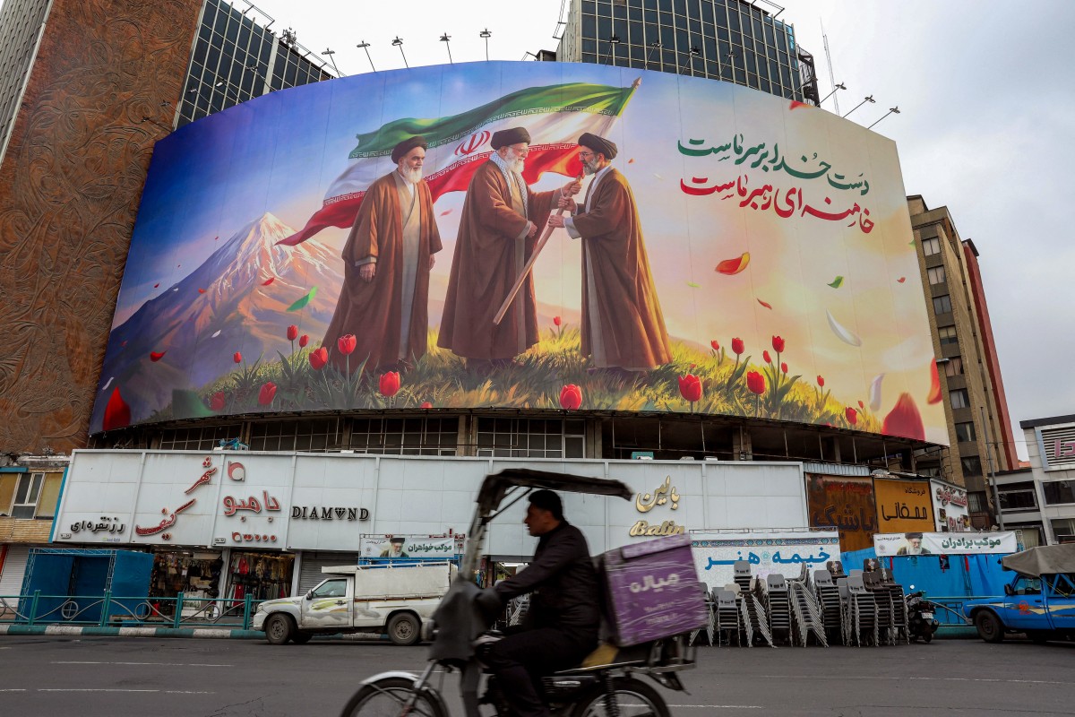 A man rides a motorcycle past a banner displayed at Valiasr Square in central Tehran on March 10, 2026, depicting Irans late supreme leader Ayatollah Ruhollah Khomeini (L) watching as his successor the late Ayatollah Ali Khamenei (C) hands over a national flag to his son and new supreme leader Mojtaba Khamenei (R).