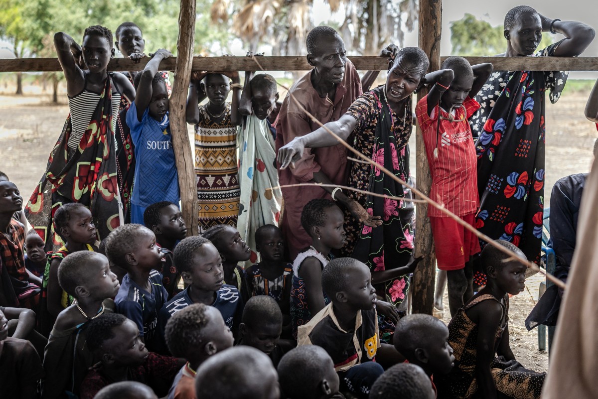 A girl from the Dinka community sits on a tree branch where people have hung their belongings as others gather under its shade at an informal site where displaced people have assembled without assistance or shelter at Yolakot informal camp near Mingkaman, on February 14, 2026. A girl from the Dinka community sits on a tree branch where people have hung their belongings as others gather under its shade at an informal site where displaced people have assembled without assistance or shelter at Yolakot informal camp near Mingkaman, on February 14, 2026.